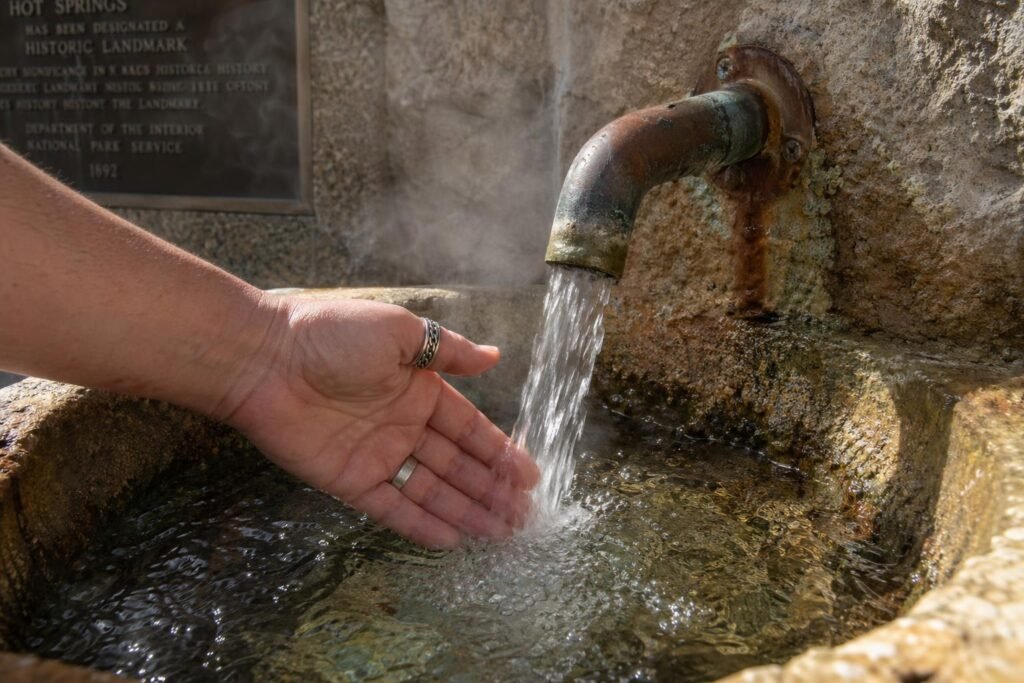Clear water pouring from a copper spigot into a stone basin, slight steam rising, a hand testing the temperature—rings visible, no polish