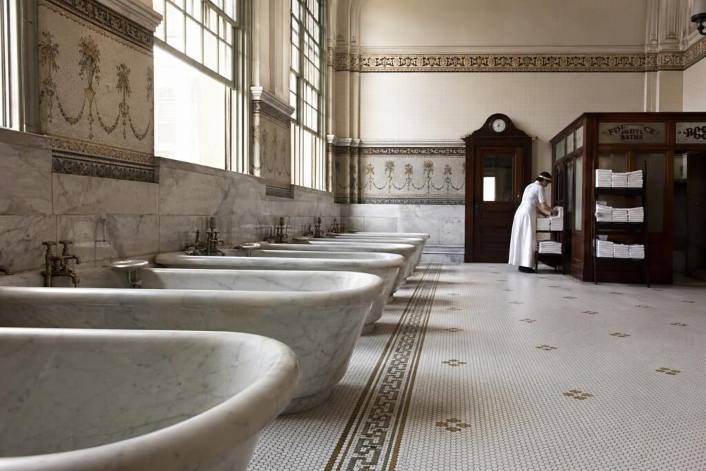 Empty white marble thermal tubs in a row, ornate tile work on walls, one attendant in period costume adjusting towels in background, completely still and quiet.