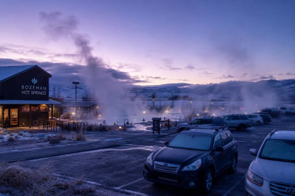 Steam clouds rising thick from outdoor pools, a few solo swimmers visible as silhouettes, parking lot half-empty with frost on car windshields