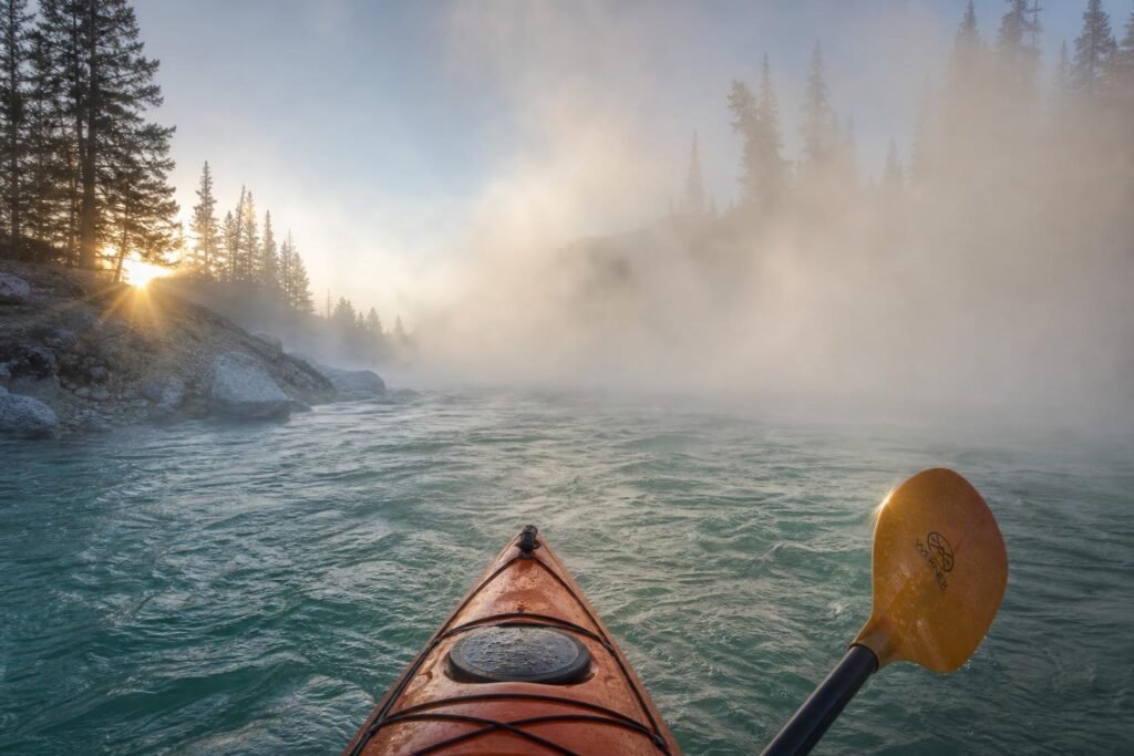 Steam rising thick off turquoise-green water with pine forest barely visible through the mist, paddle blade catching first light